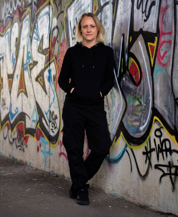 Melly – Grafikdesign & Kreativität aus dem Schwarzwald – lehnt lässig vor einer bunten Graffiti-Wand in schwarzem Hoodie und Cargohose.