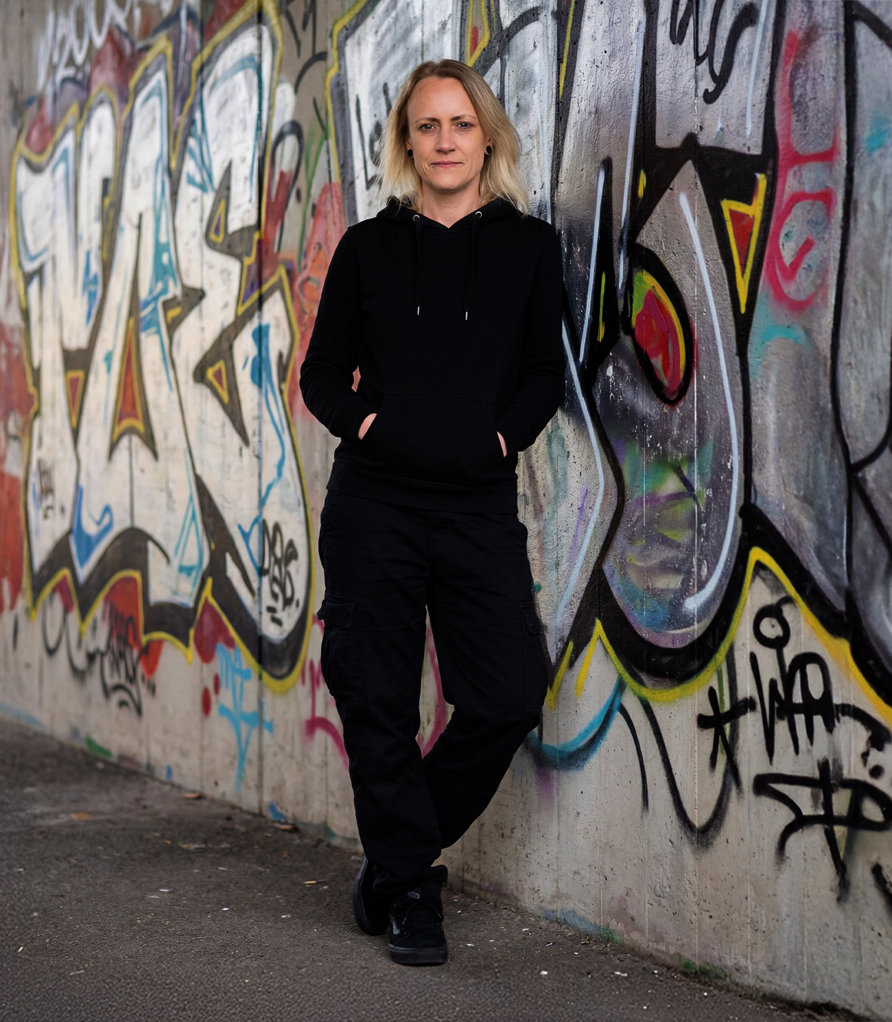 Melly – Grafikdesign & Kreativität aus dem Schwarzwald – lehnt lässig vor einer bunten Graffiti-Wand in schwarzem Hoodie und Cargohose.